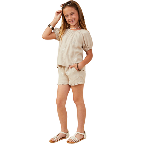 A girl wearing a beige linen-blend short set with puff sleeves, white bow embroidery, and ruffled shorts; shown from front and back.