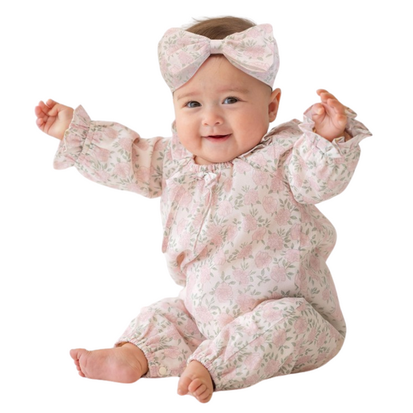 Smiling baby wearing pink floral romper and matching bow headband, sitting with arms raised and feet bare.