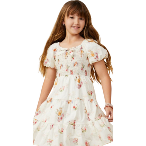Girl wearing a white smocked floral dress with puff sleeves, front bow detail, and a tiered skirt.