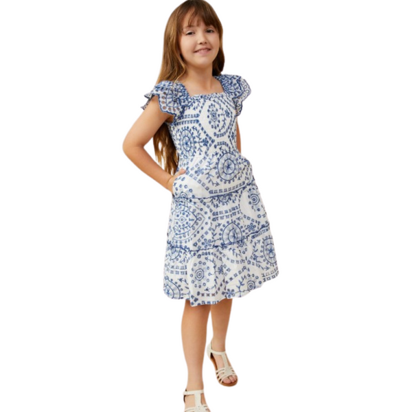 Girl in a white and blue embroidered dress, hands in pockets, showcasing a playful yet elegant profile.
