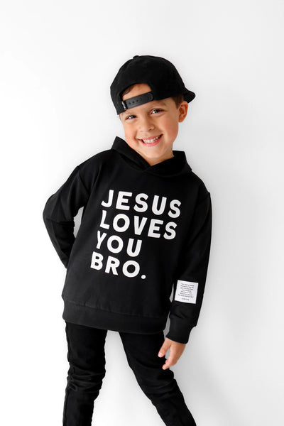 A boy modeling black hoodie with white “Jesus Loves You Bro” graphic text on front