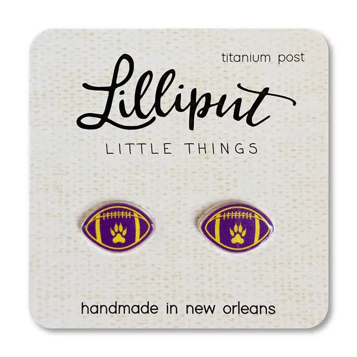 Close-up of a pair of Lilliput Little Things brand football-shaped stud earrings, featuring a purple and gold design with a paw print in the center, displayed on a white card with 'titanium post' and 'handmade in new orleans' text.