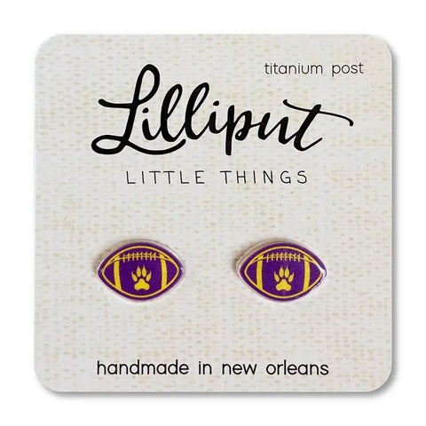 Close-up of a pair of Lilliput Little Things brand football-shaped stud earrings, featuring a purple and gold design with a paw print in the center, displayed on a white card with 'titanium post' and 'handmade in new orleans' text.