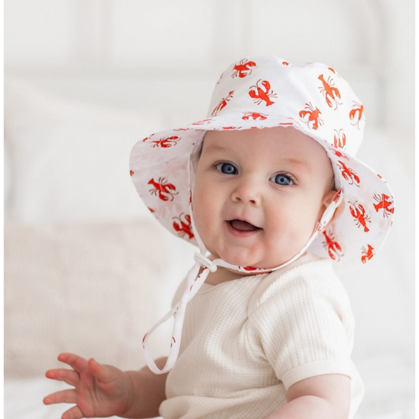 Baby wearing a white bucket hat with red lobster print, featuring a wide brim and chin strap.