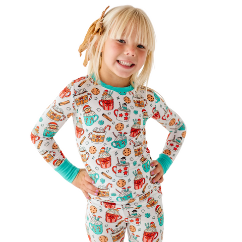 A smiling young girl with blonde hair and bangs, wearing a cute holiday-themed pajama set with a pattern of hot cocoa mugs, cookies, and candy canes, featuring teal cuffs and neckline. She has her hands on her hips and a bow in her hair.