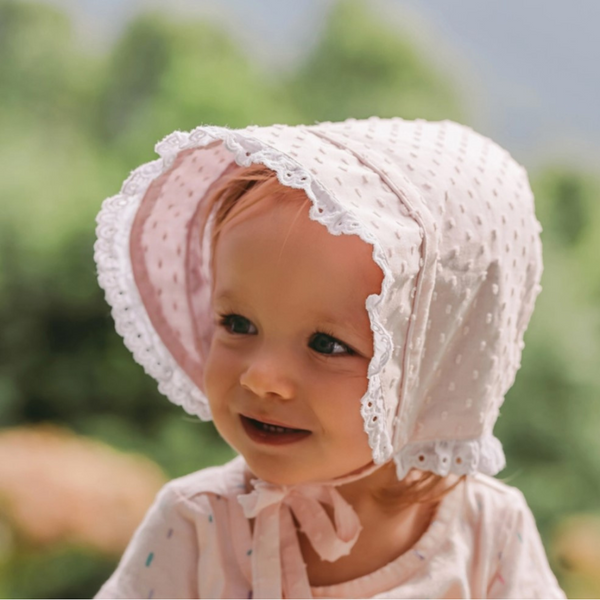 Toddler girl outdoors in a light pink clip dot bonnet with white lace trim and chin ties.