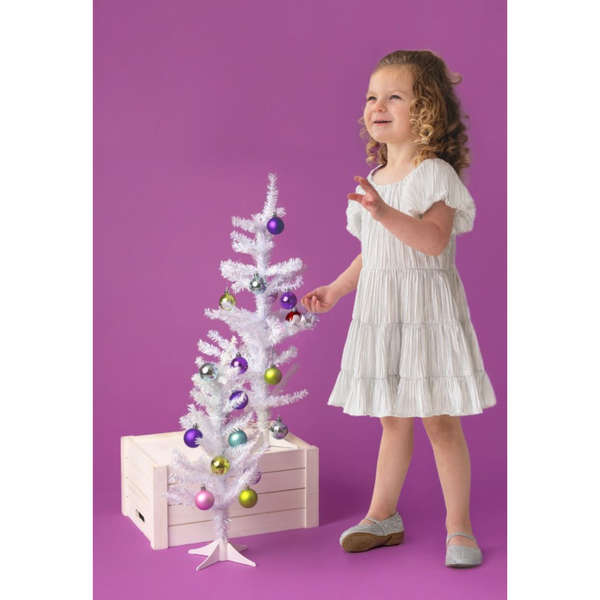 Little girl decorating a white Christmas tree, wearing a shiny silver pleated dress and sparkly shoes against a pink backdrop.

